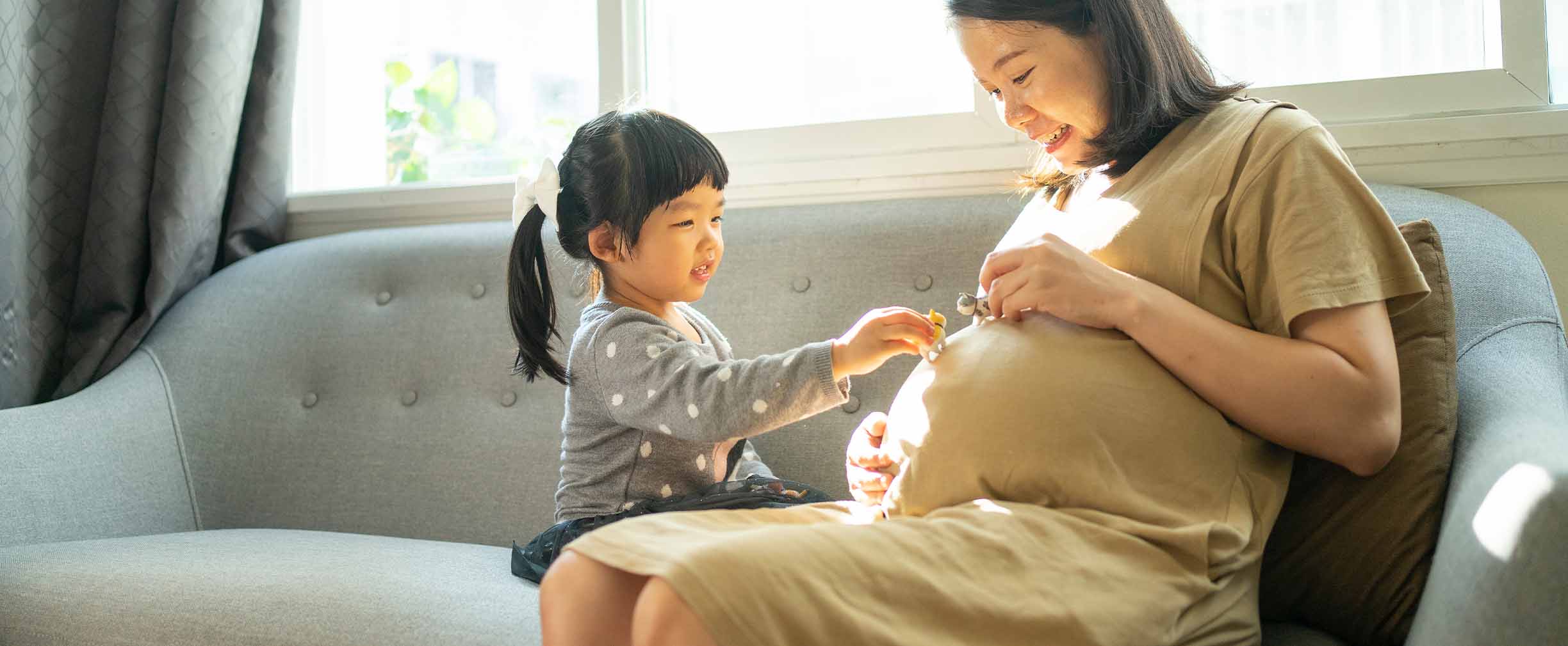 A pregnant woman plays with her young daughter on a sofa in a sunlit room