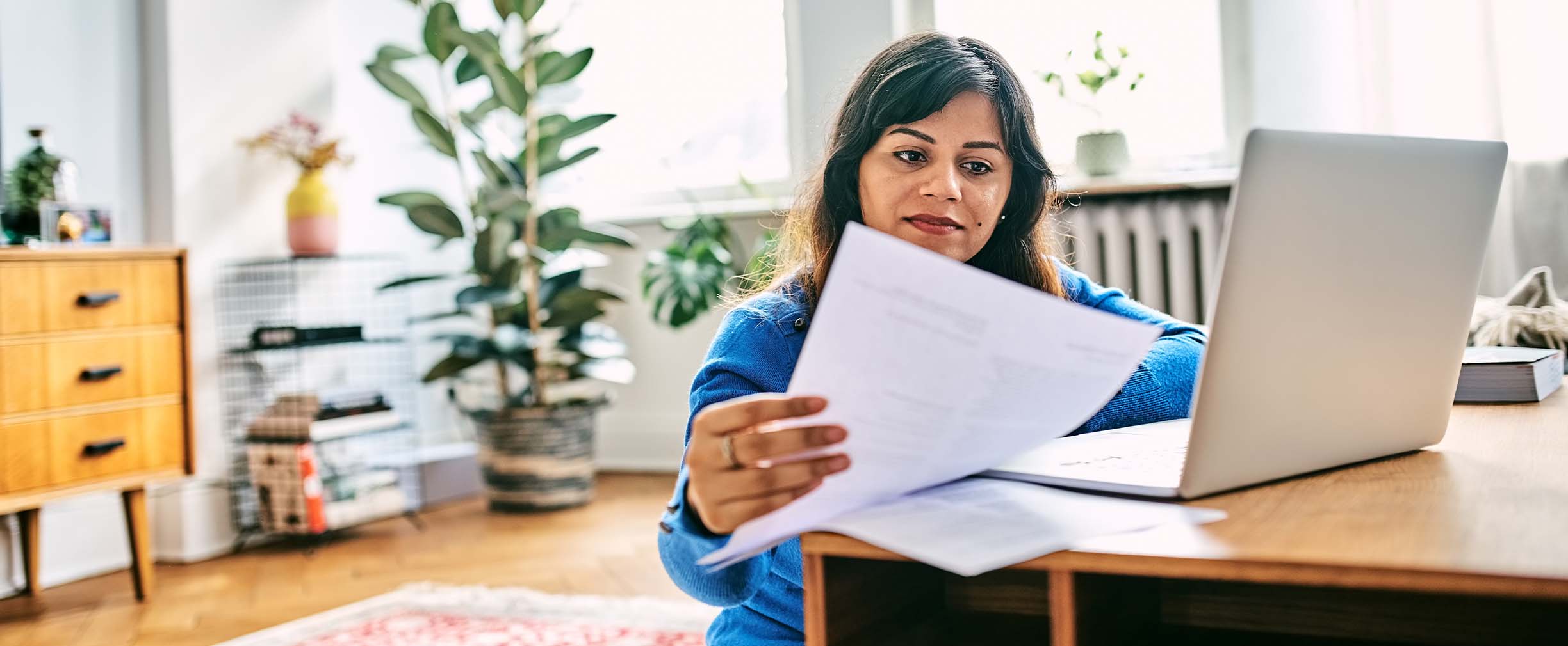 A woman reviews here finances using a laptop in a bright, plant-filled room.