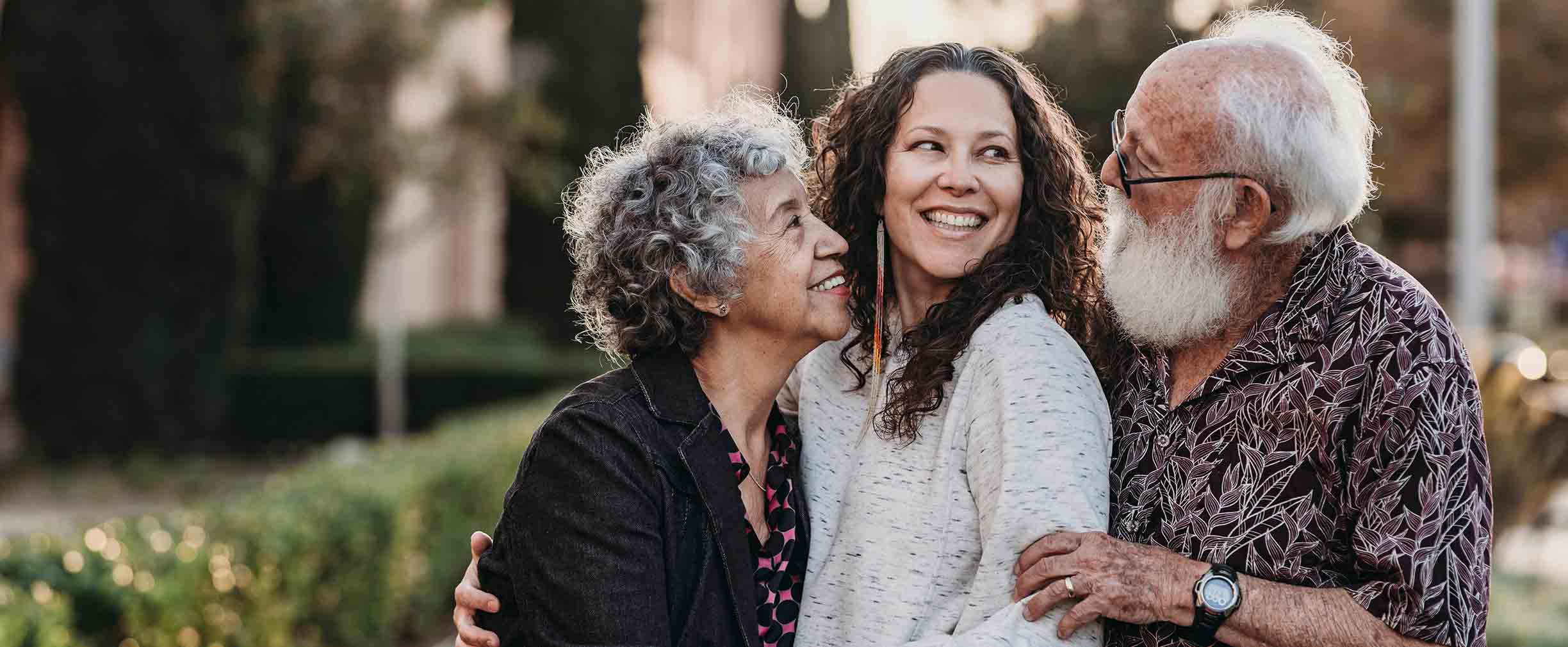 An older couple embraces their daughter in a sunny location.