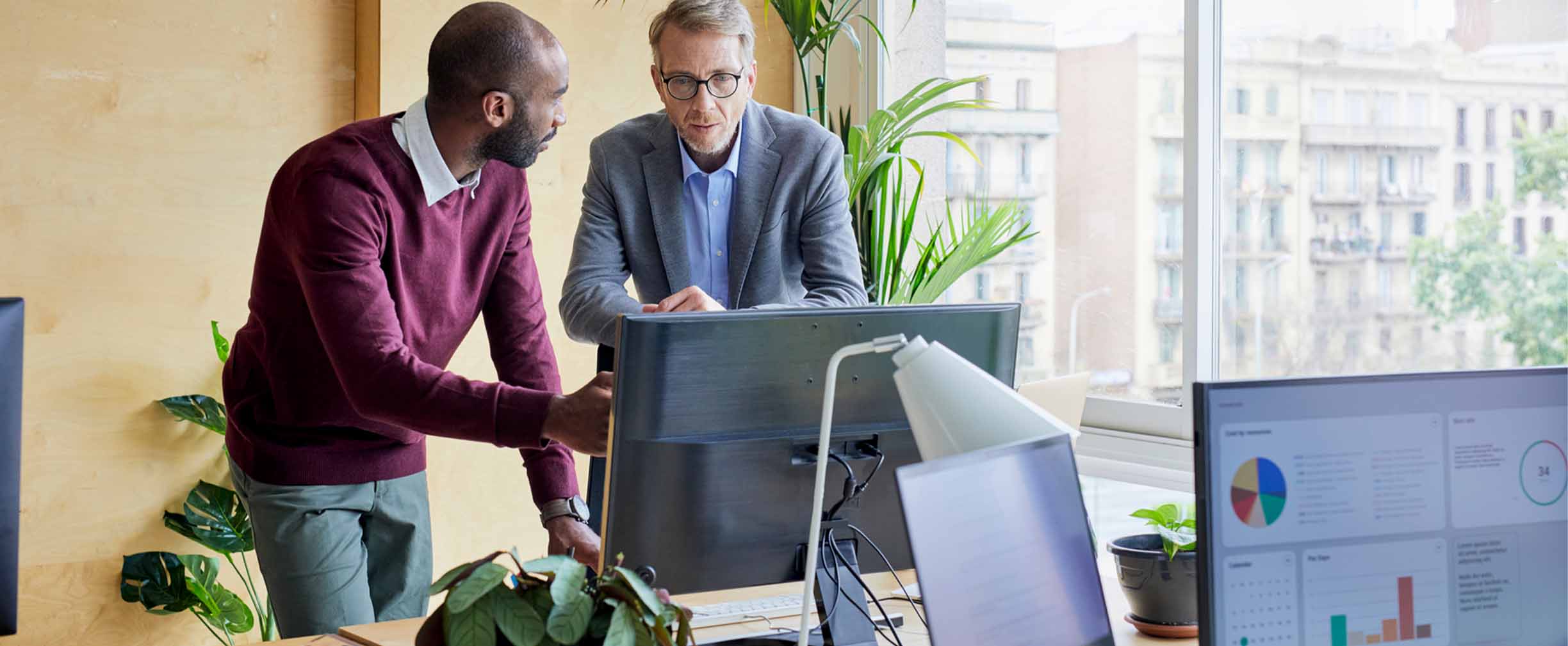 Two professionals discuss charts on a computer screen in a plant-filled office